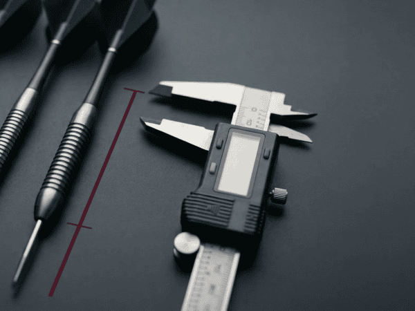 Three steel-tip tungsten darts of varying barrel thicknesses laid next to a digital precision caliper on a dark charcoal surface.