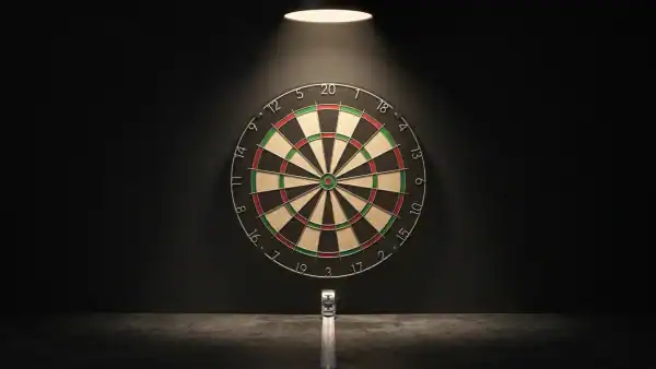 Bristle dartboard mounted on dark wall illuminated by a single overhead spotlight with a tape measure extending from the base