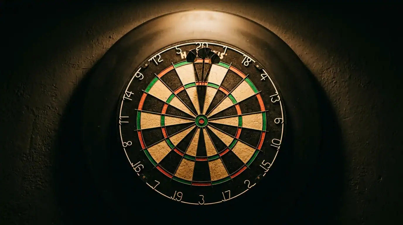 Bristle dartboard mounted on dark wall under warm spotlight with darts embedded