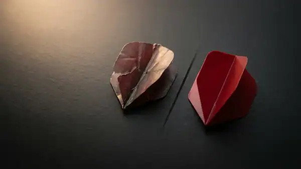 Worn torn dart flight next to a fresh crimson flight on dark charcoal surface