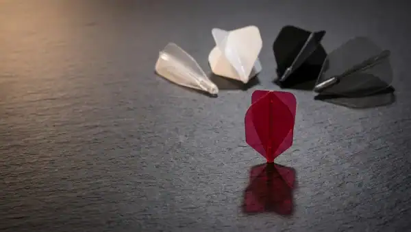 Five dart flights of different shapes arranged on dark charcoal surface with a crimson standard flight standing upright casting a geometric shadow
