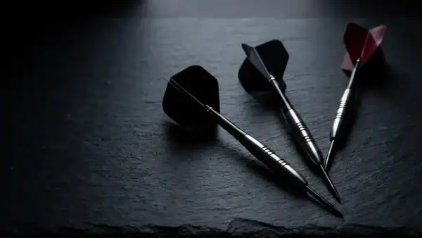 Three steel-tip tungsten darts with different flight shapes and shaft lengths arranged diagonally on a dark charcoal surface