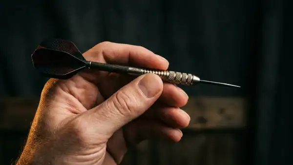 Close-up photograph of a hand holding a steel-tip dart in a pencil grip with moody lighting