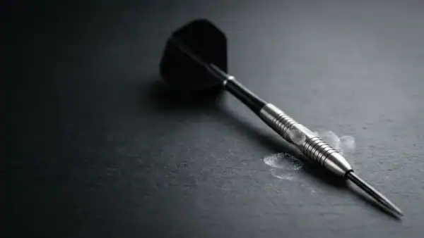 A steel-tip tungsten dart on a dark charcoal surface with subtle chalk finger marks visible on the barrel grip zone