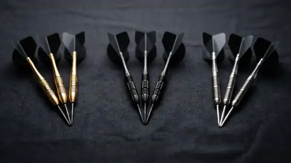 Three sets of professional tungsten darts with gold black and silver barrel finishes on dark charcoal surface