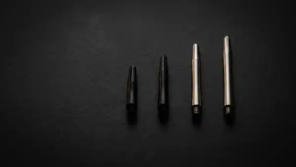 Four dart shafts arranged by length on dark charcoal surface showing extra-short and short black nylon next to medium and long polished silver aluminium