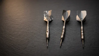 Three tungsten darts on a dark slate surface, one with crumpled flights representing common darts mistakes